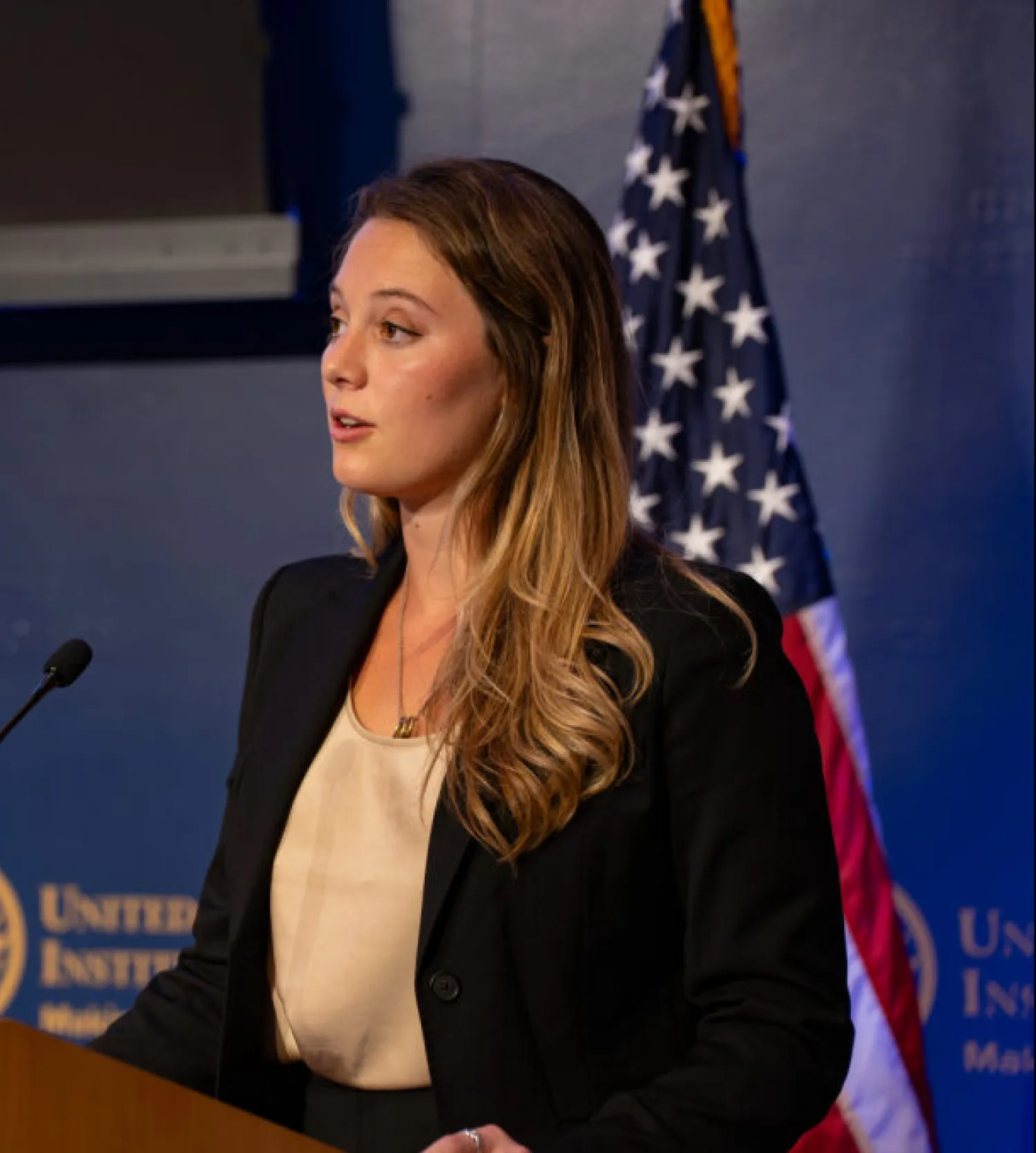 Side image of a woman standing at a lectern with the US flag in the background