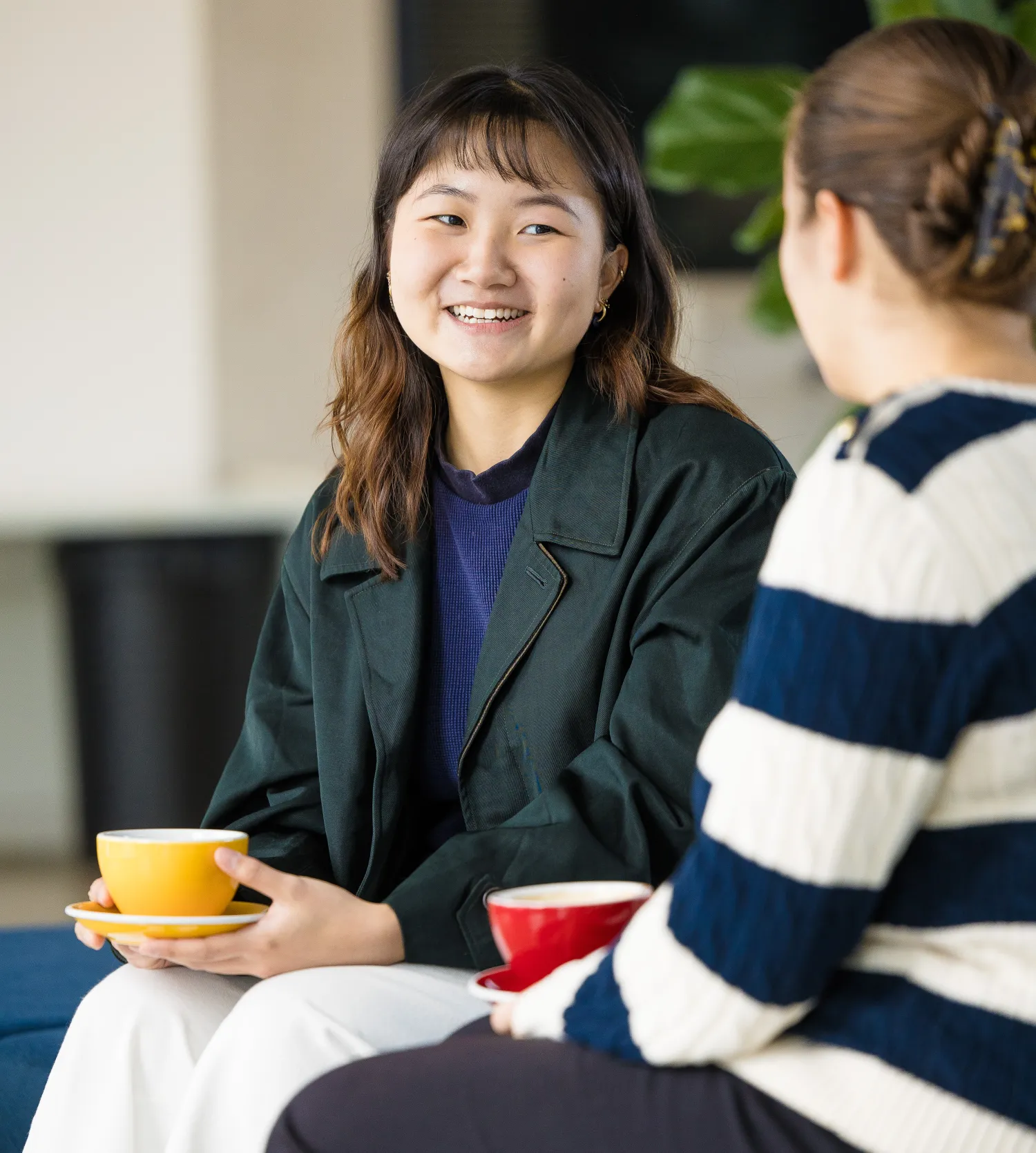 image of two female students sat by one another, one with their back to the camera, holding coloured coffee cups