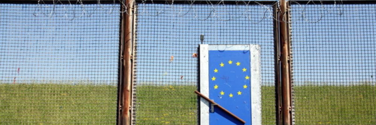 Image of a tall barbed wire fence with a door in with the EU symbol on. 