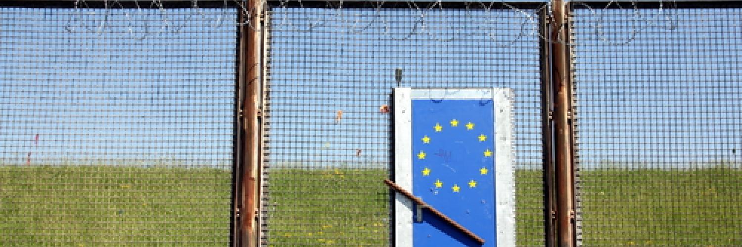 Image of a tall barbed wire fence with a door in with the EU symbol on. 