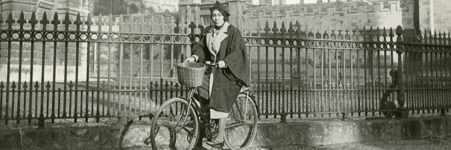 Image of a woman sat on a bicycle in front of a fence and a building