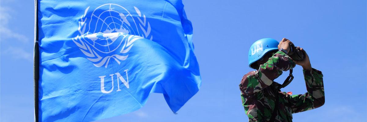 UN Peacekeeper looking out with binoculars, UN flag on the left, blue sky background