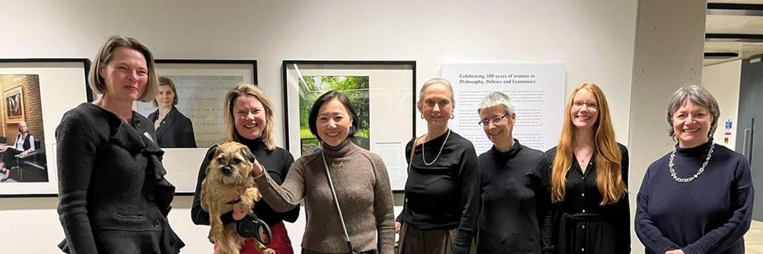 Seven women smiling at the camera, stood in front of some exhibition photographs and text.