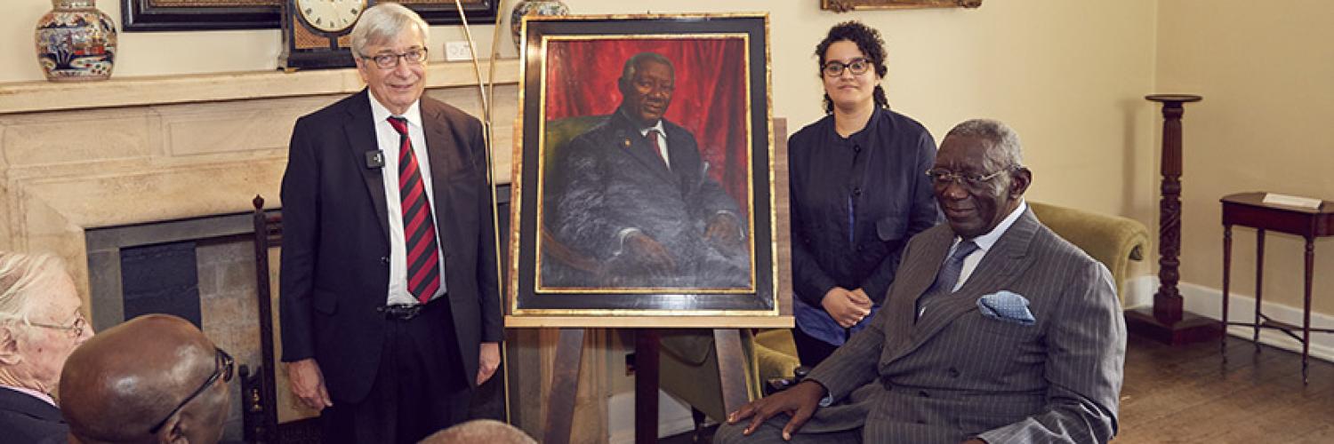 Two people stand and one sits aside a painted portrait in a room, in front of an audience