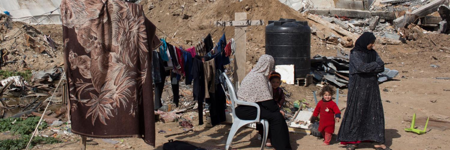 Two women and a child sit and stand by some washing, drying on a line, in front of war torn buildings