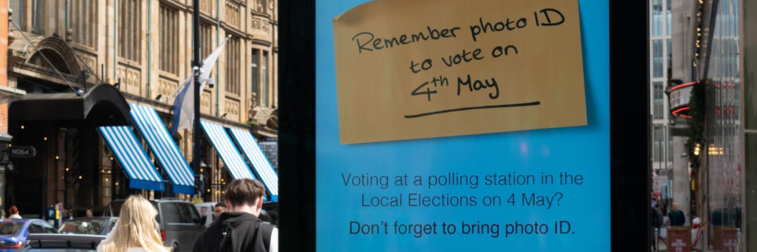 Two people walk past an electronic street sign advertising 'Remember photo ID to vote on 4 May' message from the UK Electoral Commission. 