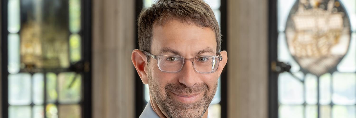 Image of a man in front of a window wearing a tie and shirt and glasses smiling at camera