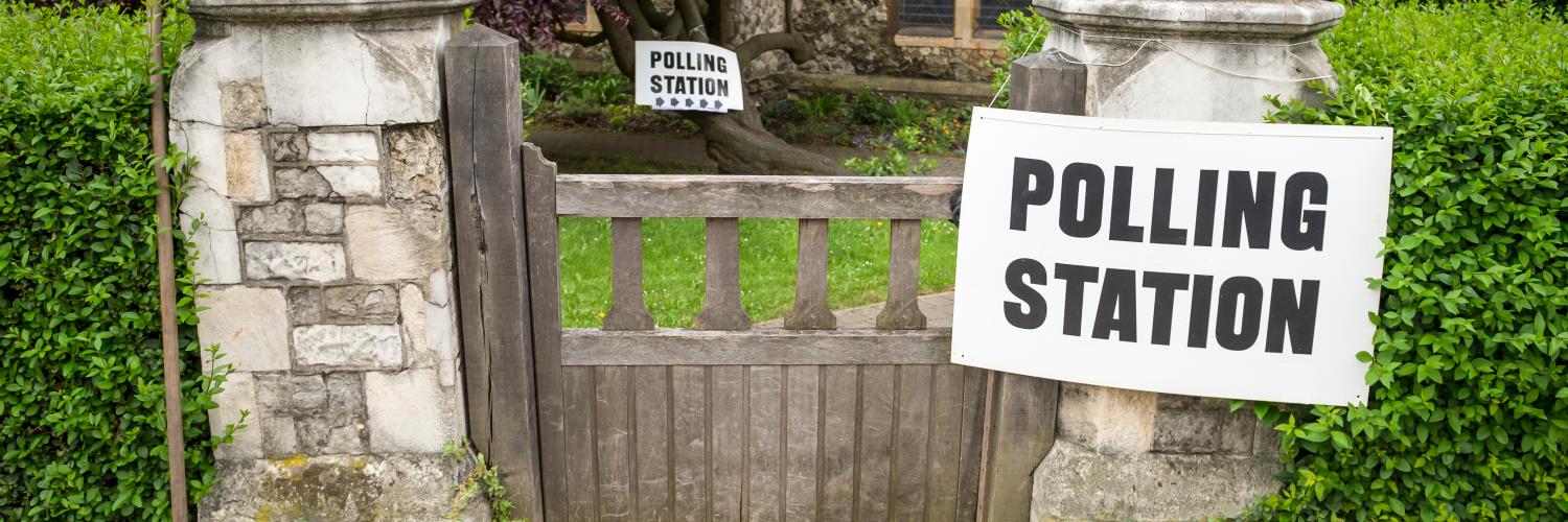 Photograph of an entrance gate to a polling station marked by two white signs with bold black text reading "POLLING STATION." 