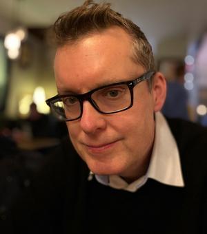 Image of a man wearing glasses and a dark jumper and white shirt looking at the camera