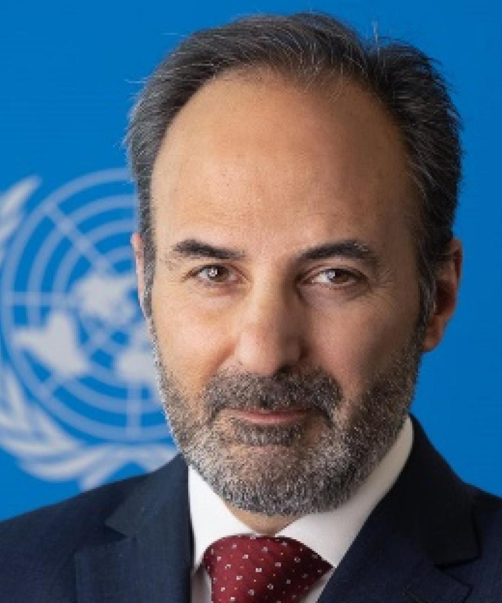 Image of a man in front of a blue screen with the UN logo behind him smiling at the camera