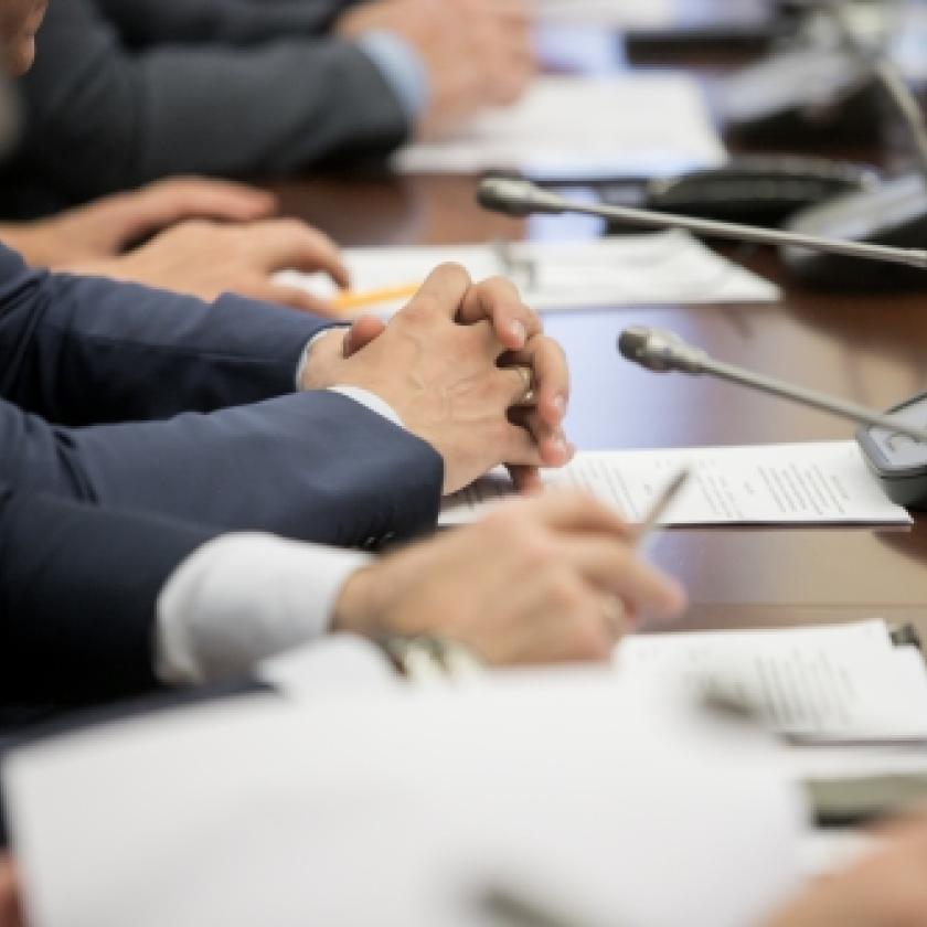 A group of politicians sit at a table with papers and microphones during a meeting.