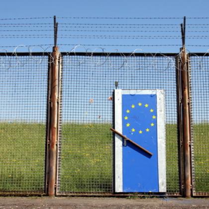 Image of a tall barbed wire fence with a door in with the EU symbol on. 