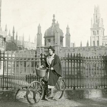 Image of a woman sat on a bicycle in front of a fence and a building