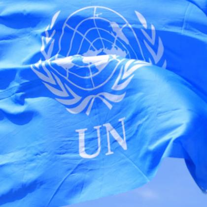 UN Peacekeeper looking out with binoculars, UN flag on the left, blue sky background