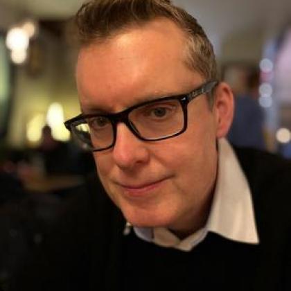 Image of a man wearing glasses and a dark jumper and white shirt looking at the camera
