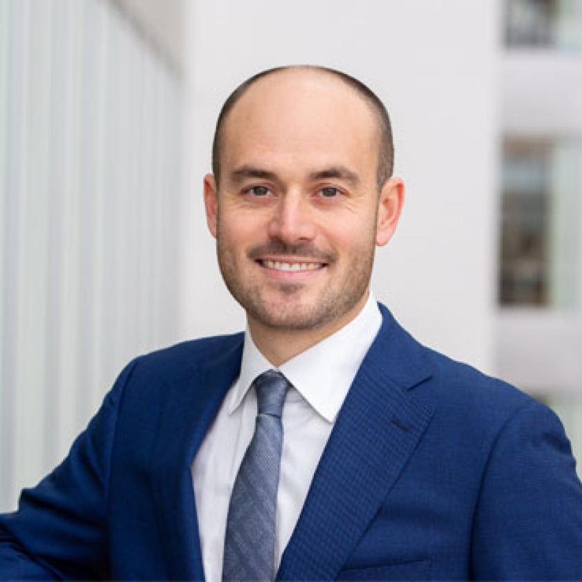 Image of a man smiling at camera wearing a blue suit and tie with a building behind him