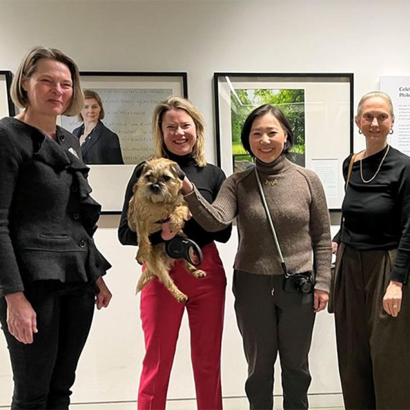 Seven women smiling at the camera, stood in front of some exhibition photographs and text.