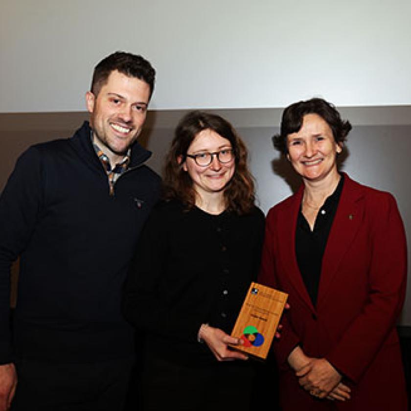 Image of a woman holding a certificate stood in front of a wall with a man on one side of her and a woman on the other side of her, with the University of Oxford logo on the wall