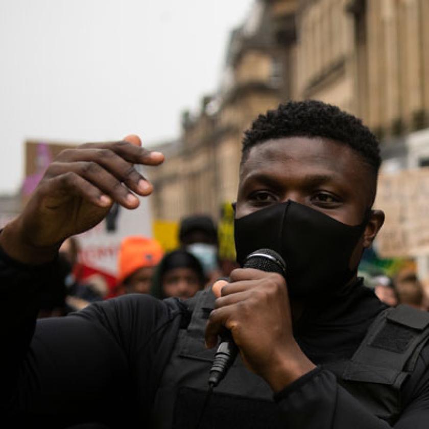 Image of a black man facing camera with a microphone in his hand with a policeman facing him