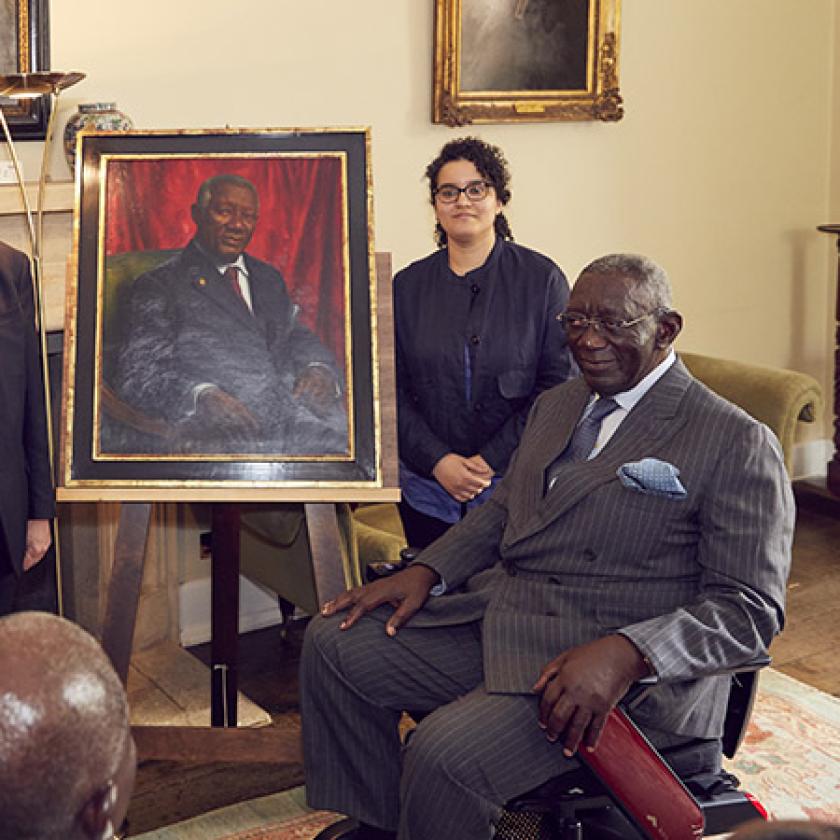 Two people stand and one sits aside a painted portrait in a room, in front of an audience