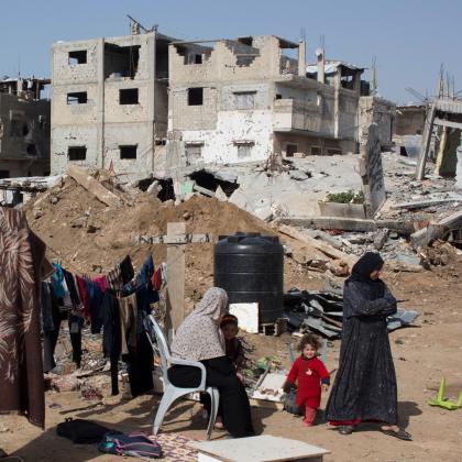 Two women and a child sit and stand by some washing, drying on a line, in front of war torn buildings