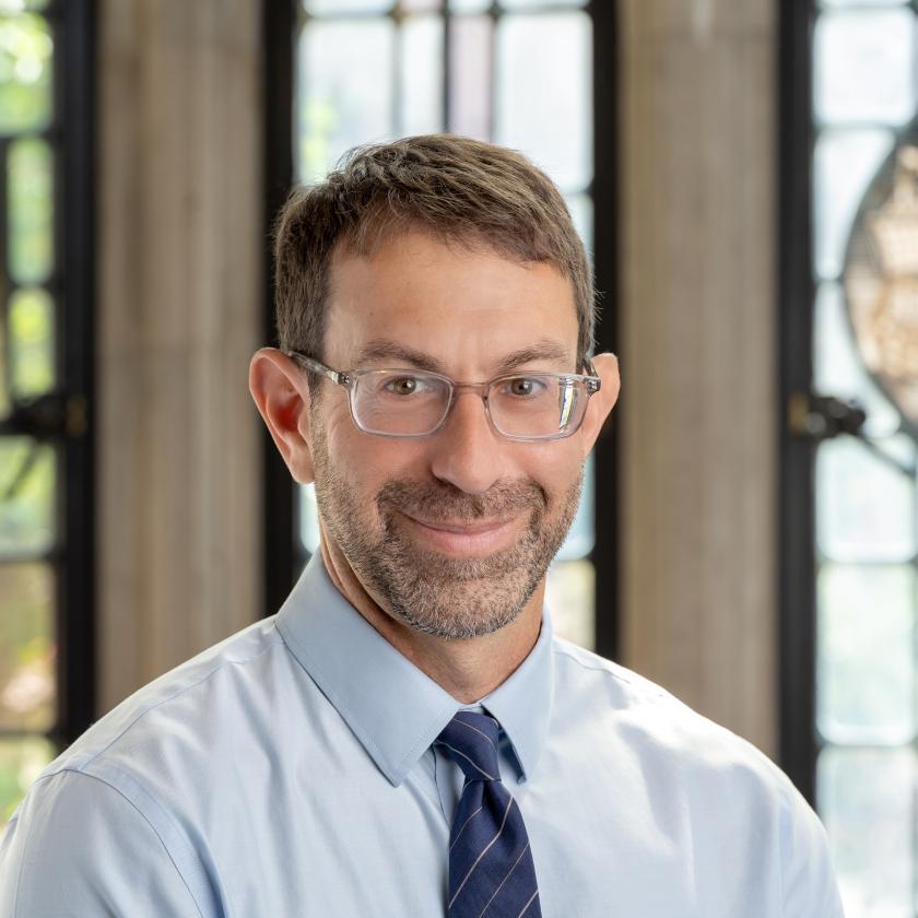 Image of a man in front of a window wearing a tie and shirt and glasses smiling at camera