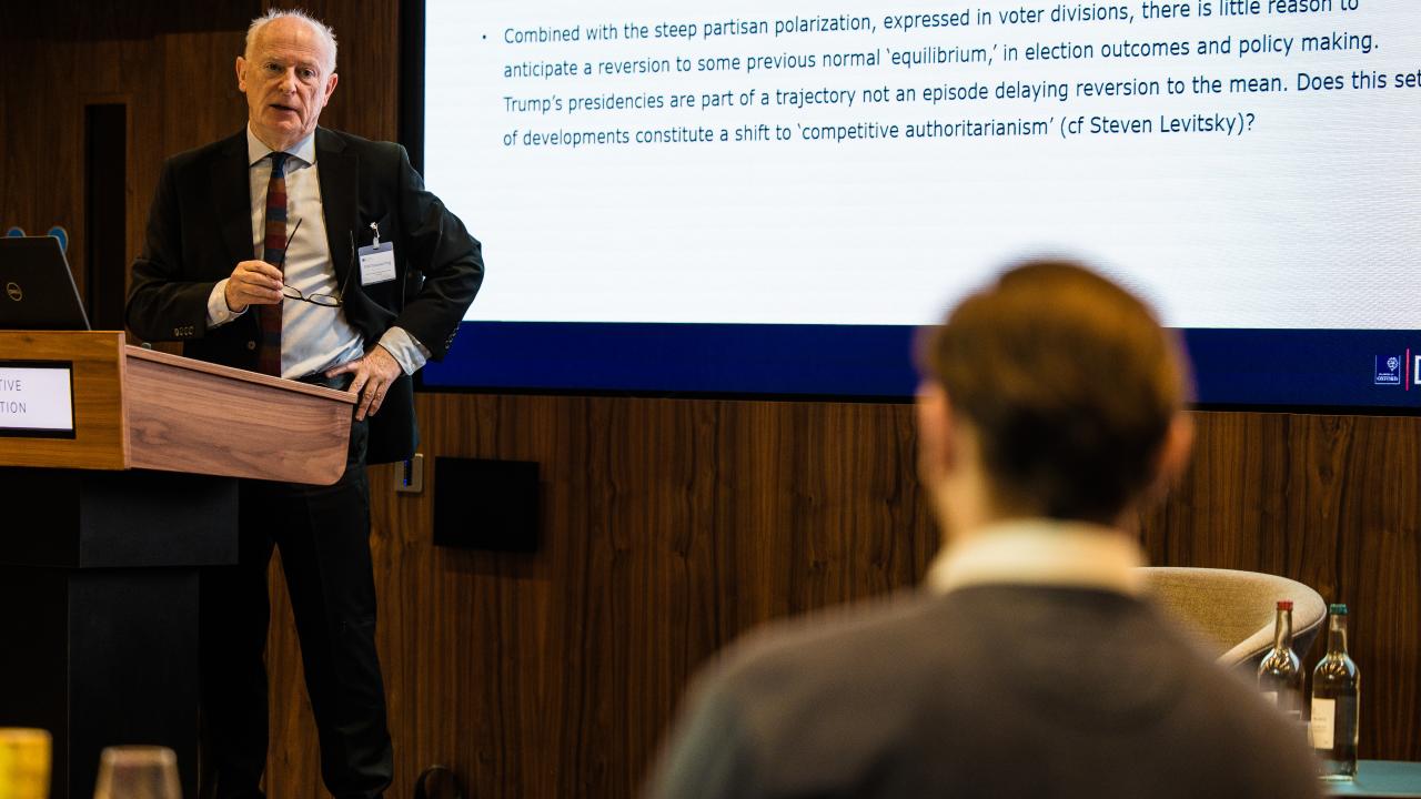 image of a man stood at a lectern giving a lecture with a man with his back to the camera in the audience