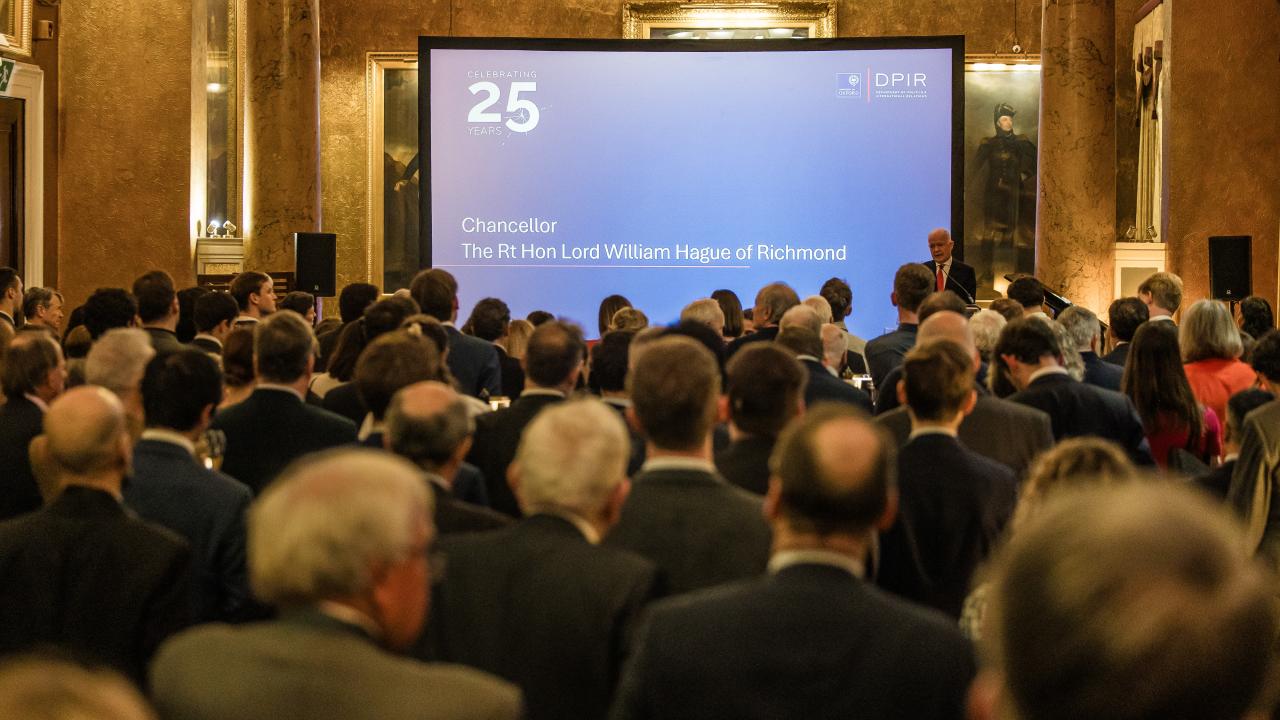 image from behind of a seated audience in a large room in front of a projector screen