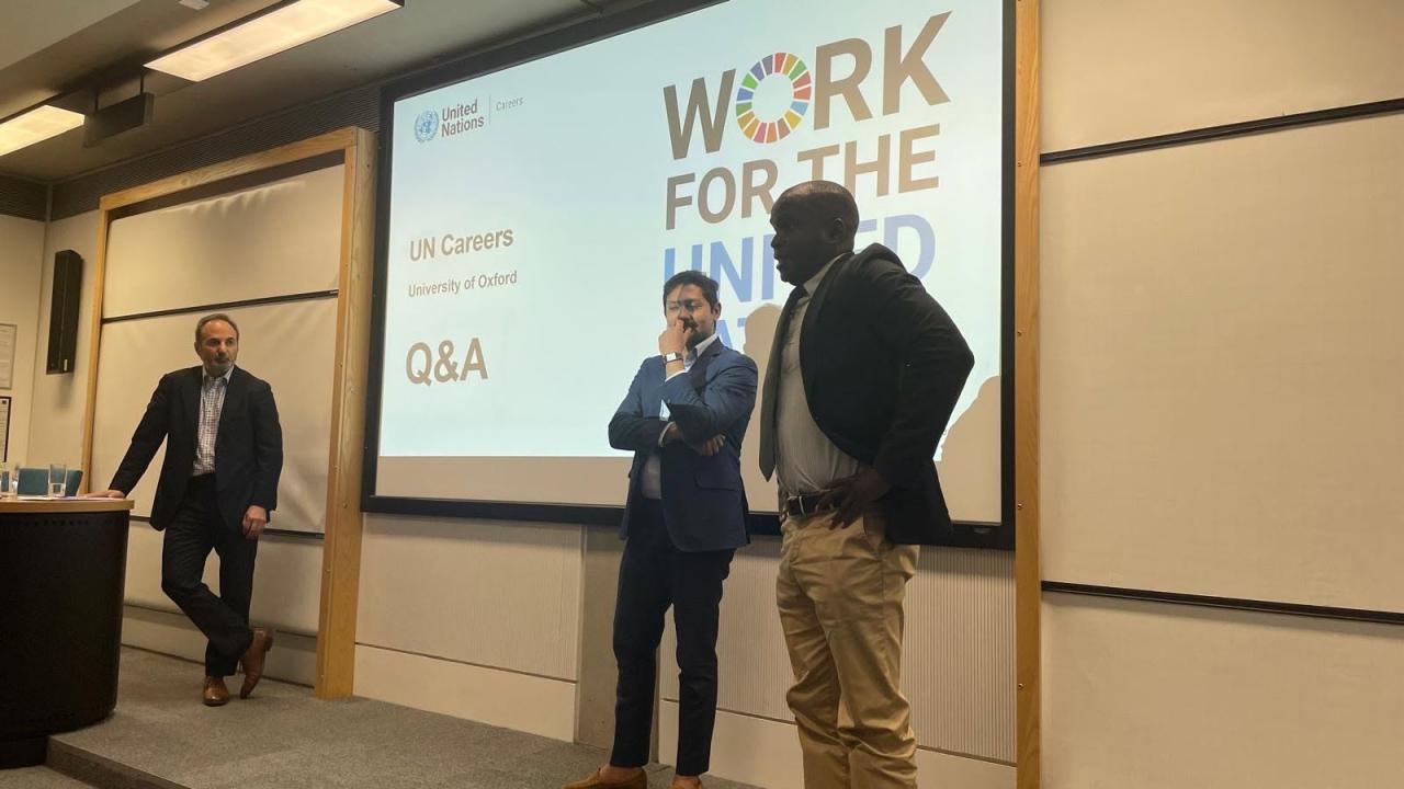 image of two men stood in front of a screen with the words 'Work for the United Nations' on it