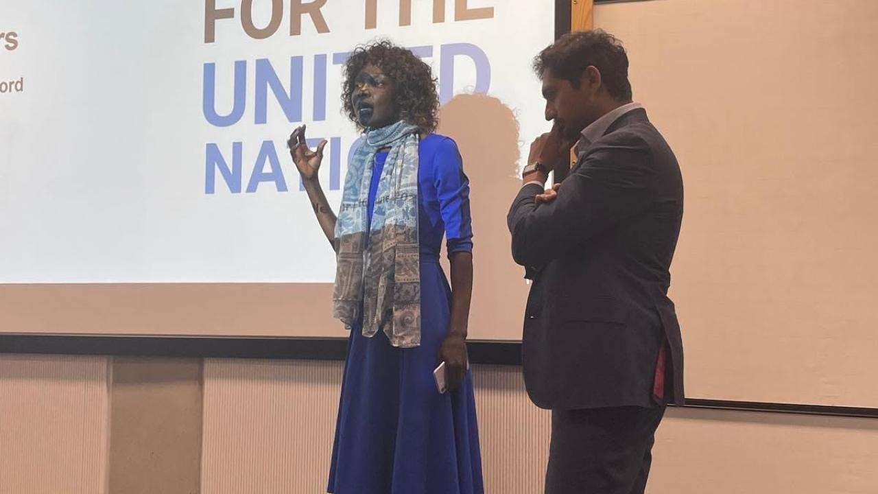 image of a man and woman stood in front of a screen with the words 'Work for the United Nations' on it