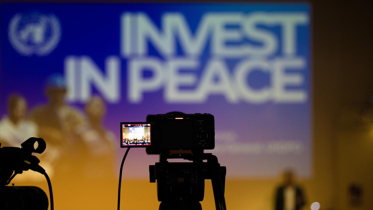 image of the silhouette of a camera in front of a screen with the words 'Invest in Peace' on it