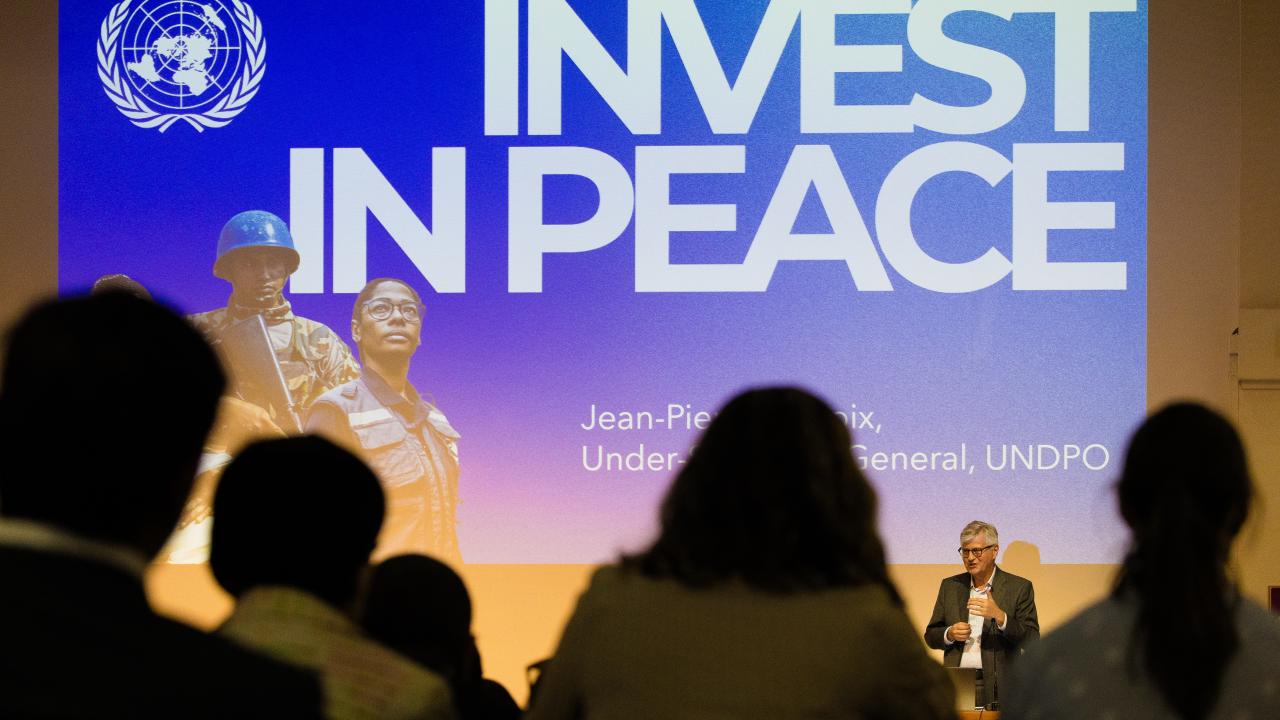 image of the rear of an audience in front of a screen with the words 'Invest in Peace' on it