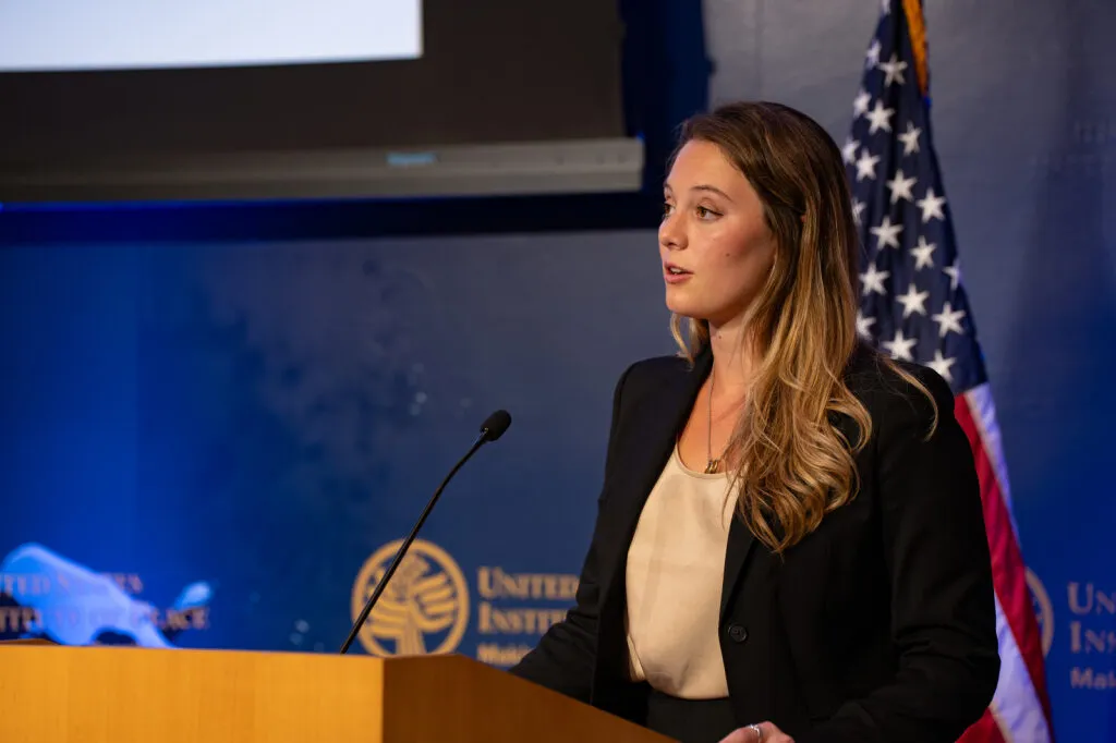 Side image of a woman standing at a lectern with the US flag in the background