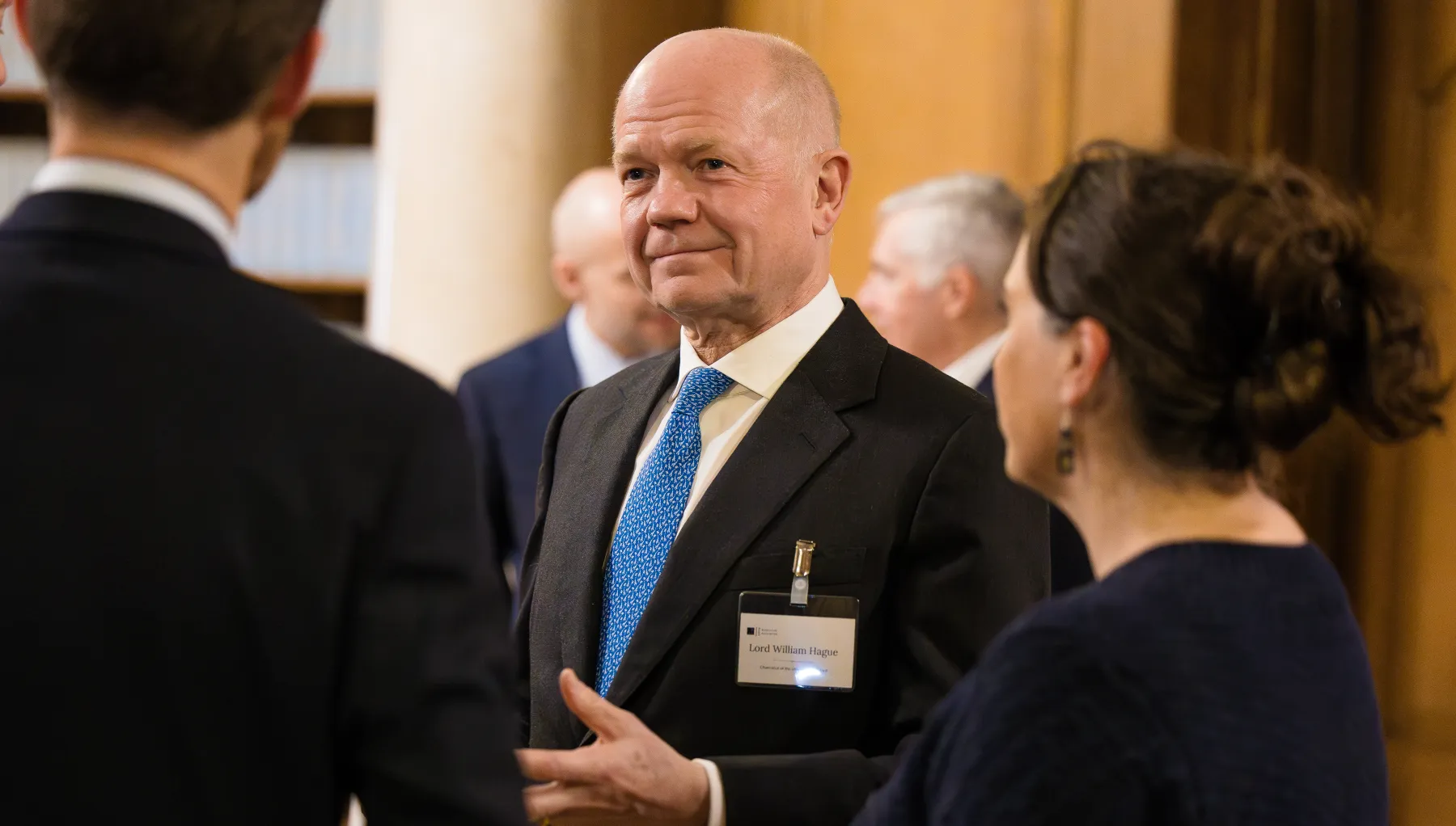 Image of a person in a suit and tie talking to two people with their backs to the camera