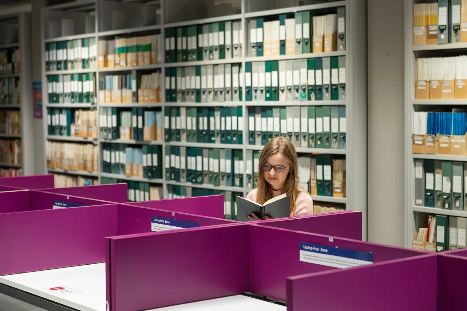 Young woman reading a book in a no laptop zone, with library shelves in the background 