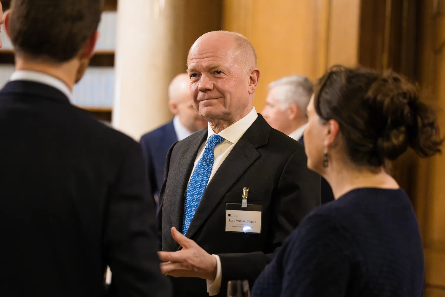 Image of a person in a suit and tie talking to two people with their backs to the camera