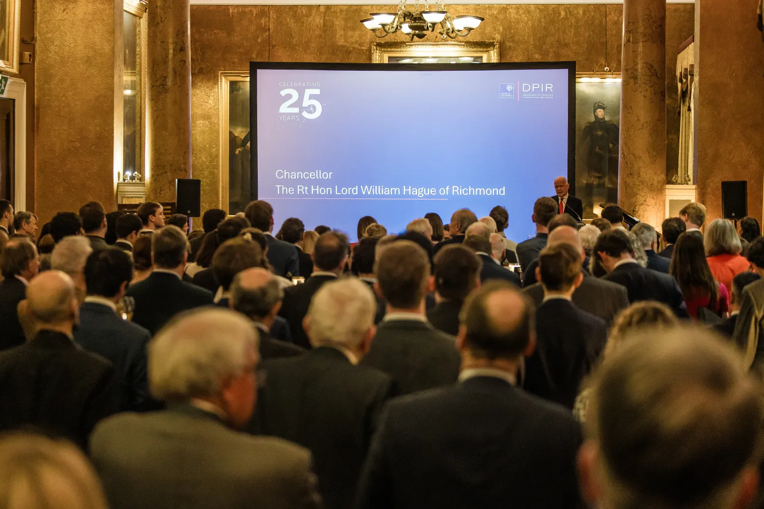 image from behind of a seated audience in a large room in front of a projector screen