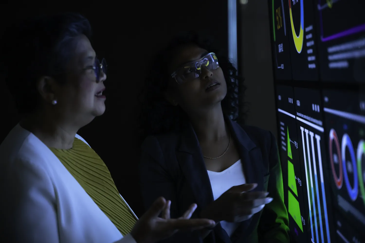 Two women analysing complex data charts on large futuristic digital screen