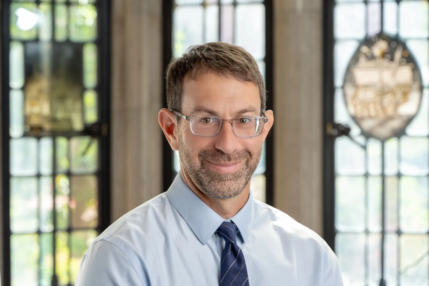 Image of a man in front of a window wearing a tie and shirt and glasses smiling at camera