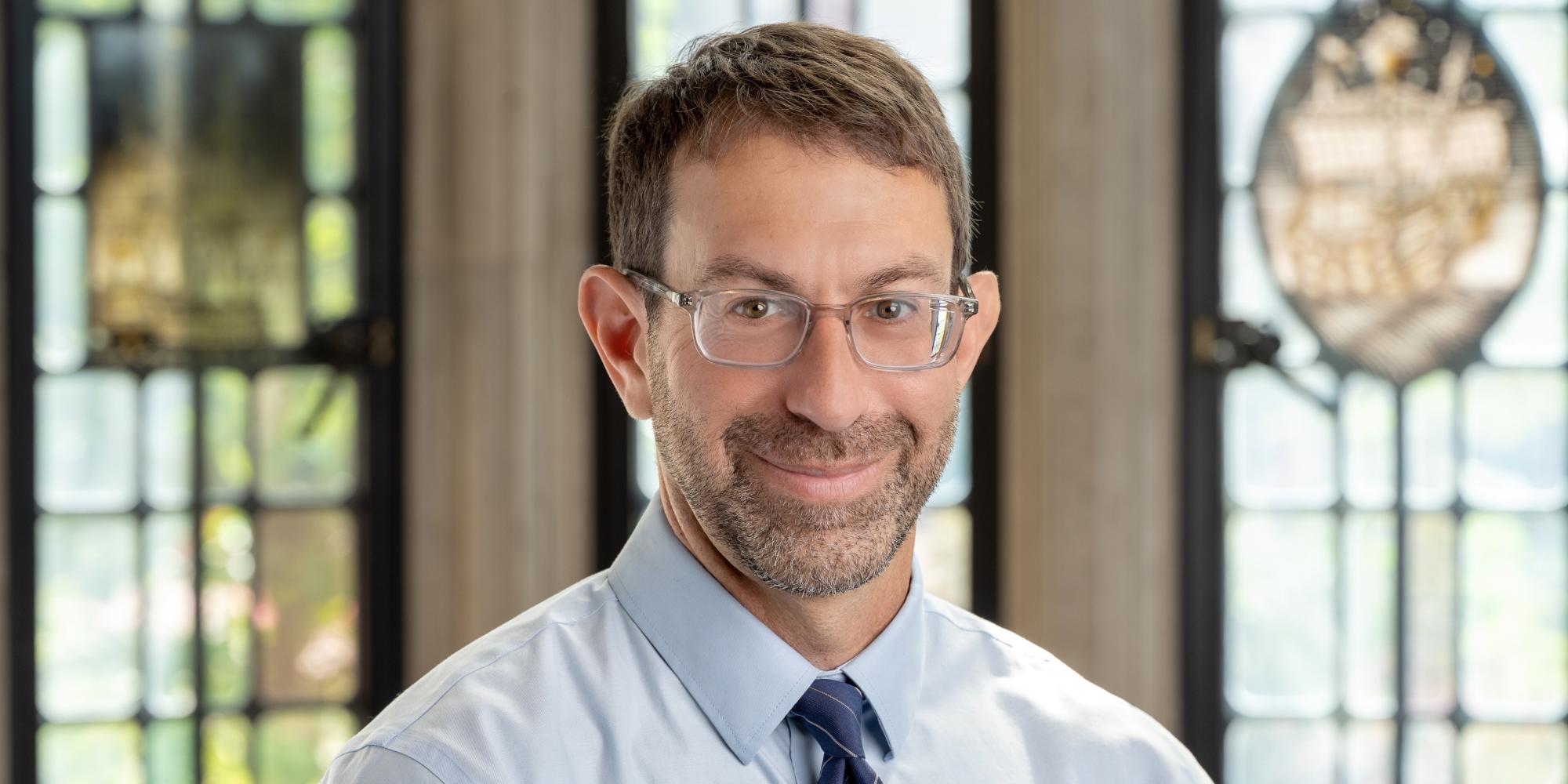 Image of a man in front of a window wearing a tie and shirt and glasses smiling at camera