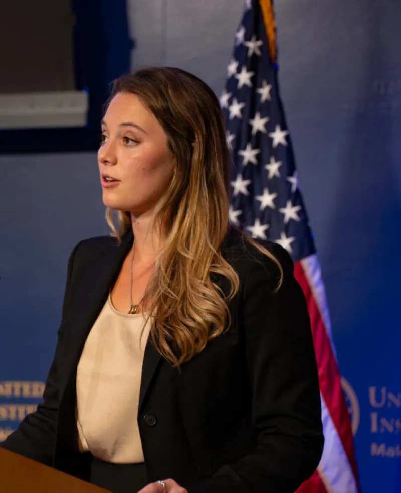 Side image of a woman standing at a lectern with the US flag in the background